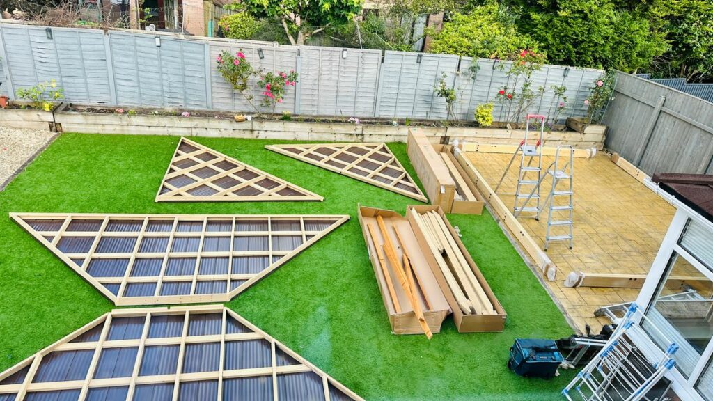 An overhead view captures the unboxing and preparation phase for assembling a large wooden outdoor structure, possibly a gazebo, pergola, or pavilion, in a suburban backyard.

Materials and Components
The components of the structure are laid out on a vibrant artificial grass lawn.

Roof/Wall Panels: Four large, triangular panels are positioned in the foreground and center.

Two smaller triangles feature a light wood lattice design.

Two larger, wider triangles are composed of a light wood frame with a dark brown, corrugated material (likely polycarbonate or metal roofing), suggesting they are roof sections.

Structural Timber: Two open cardboard boxes contain various sizes of light-colored wooden beams and posts, which are the main structural elements of the kit.

Packaging: The rest of the disassembled structure's parts are contained within long, rectangular cardboard boxes situated near the back and right side of the lawn.

Tools/Equipment: A small black tool bag or box is visible near the bottom center, and a silver stepladder is set up on the patio area, indicating active construction.

Backyard Setting
The assembly is taking place in a neat and well-maintained residential garden:

Ground Cover: The garden is split between the artificial green grass and a paved area or patio composed of lighter, square pavers on the right.

Fencing: The background is lined with a light gray wooden fence.

Landscaping: A raised garden bed runs along the base of the fence, featuring some soil, a few decorative elements, and climbing pink roses growing against the fence.

Adjacent Structures: A small section of what appears to be a white conservatory or garden room is visible in the bottom right corner, reflecting the light.

The photograph documents the initial, organised stage of a DIY outdoor construction project.