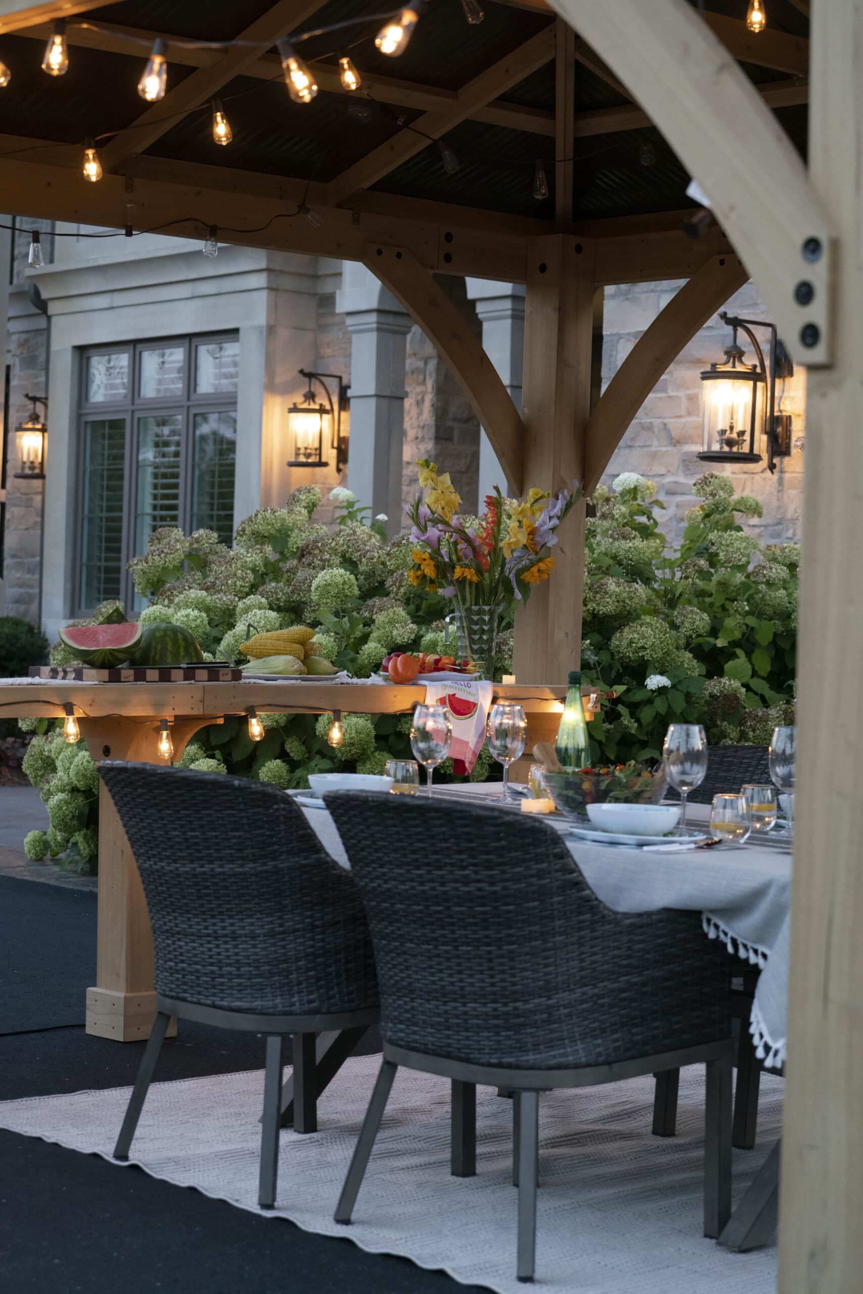Outdoor dining setup under a wooden pergola with string lights, featuring a white tablecloth, wicker chairs, and a fruit-laden counter beside a stone house and hydrangea bushes.