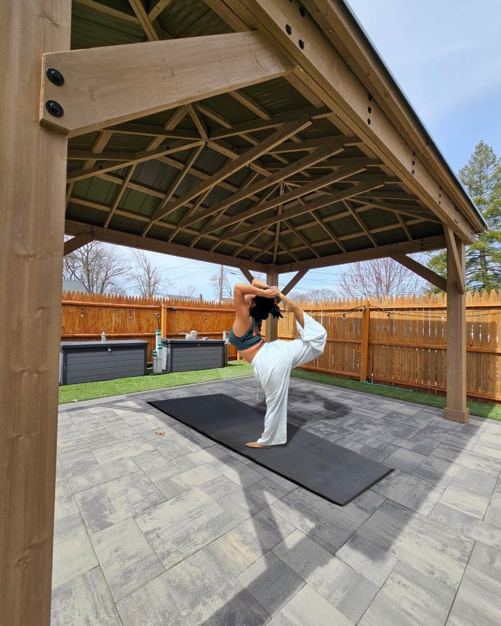 This image captures a woman performing a yoga pose, specifically the Natarajasana (Dancer's Pose) or a variation of it, beneath a wooden outdoor pavilion or gazebo.

The woman is centered on a black yoga mat laid out on a gray paver patio. She is standing on her left leg, with her right leg extended upwards and bent at the knee, holding her right foot with her right hand behind her back. She is leaning forward, maintaining balance and deep extension. She is wearing a dark sports top and loose-fitting white yoga pants.

The pavilion structure above her has a solid, light brown wood frame, with large, exposed beams and metal fasteners visible in the foreground. The roof's underside reveals a complex dark-colored metal frame with a patterned support structure. The shelter provides an ideal, shaded space for outdoor practice.

The surrounding area consists of a neatly laid, large-format gray paver patio. A section of synthetic turf or grass is visible just beyond the pavers. The yard is enclosed by a light brown wooden privacy fence. Against the fence, two large, dark gray storage bins or containers are placed. The setting is a clean, modern backyard on a clear, bright day, emphasising the integration of wellness into the outdoor living space.