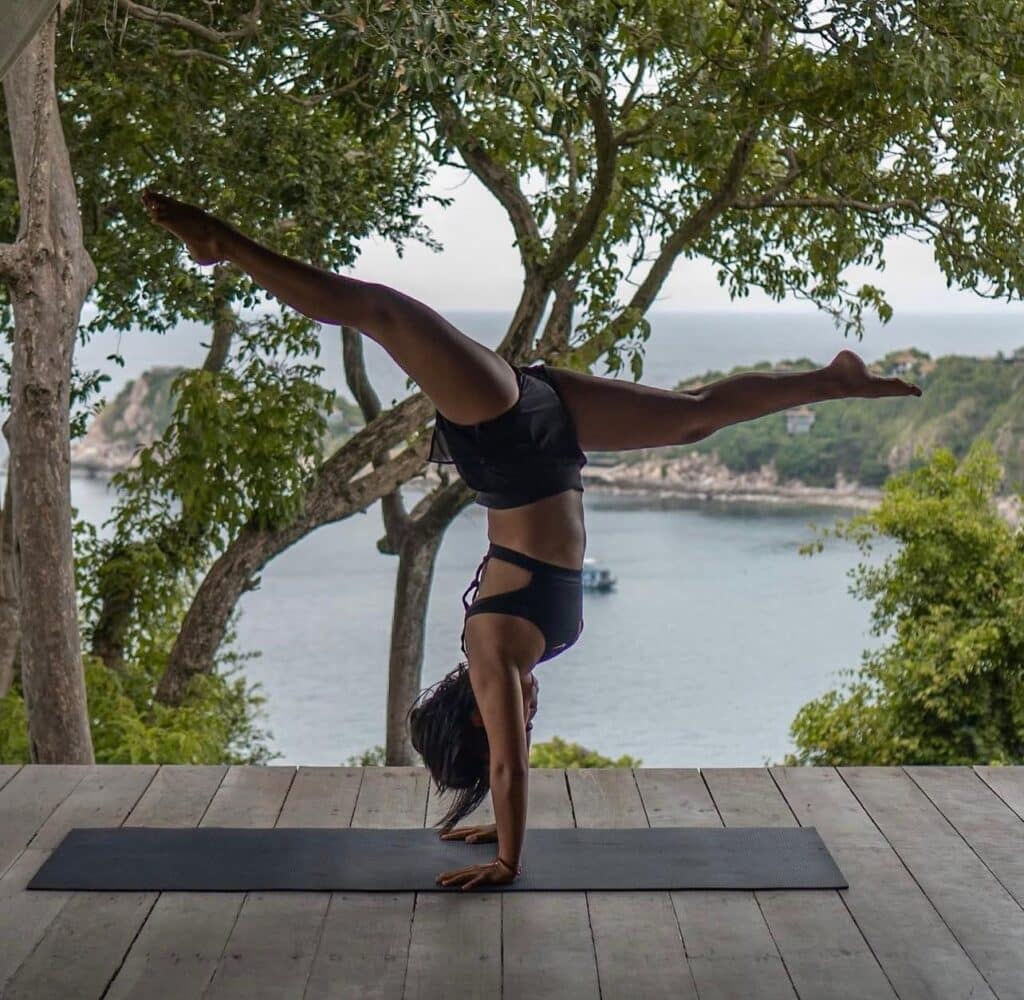 This image features a woman demonstrating an advanced yoga or acrobatic pose—a handstand split—on a wooden deck overlooking a tropical coastline.

The woman is centered in the frame, wearing a black athletic outfit consisting of a sports bra and black shorts. She is in a perfectly vertical handstand position, supported by her hands planted on a dark yoga mat laid out on the light-colored wooden deck floor. Her legs are extended into a full split, with one leg reaching high above her head and the other stretched parallel to the deck, creating a striking line that spans the width of the image. This demonstrates significant strength, balance, and flexibility.

The wooden deck forms the foreground, and the setting appears to be a raised platform or balcony, possibly part of a yoga retreat or resort. The background offers a stunning, natural view: framing the central figure are lush green trees and foliage, providing natural shade. Beyond the trees, a wide expanse of deep blue-gray ocean is visible, with a small rocky cove and a distant island or headland jutting into the water. A small boat is anchored in the bay. The composition places the dynamic human figure against a serene, natural, and exotic coastal landscape.