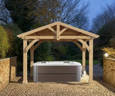 Wooden pergola with a gabled roof and exposed beams built over a modern rectangular hot tub with a gray exterior and white interior, set on a cobblestone patio between brick walls, with trees and shrubbery in the background during twilight.