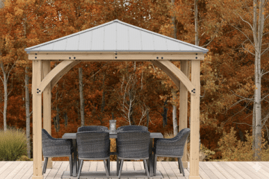 Wooden gazebo with a metal roof, featuring a rectangular dining table with six woven-style chairs on a wooden deck, surrounded by autumn trees with orange and brown foliage in a forest setting.