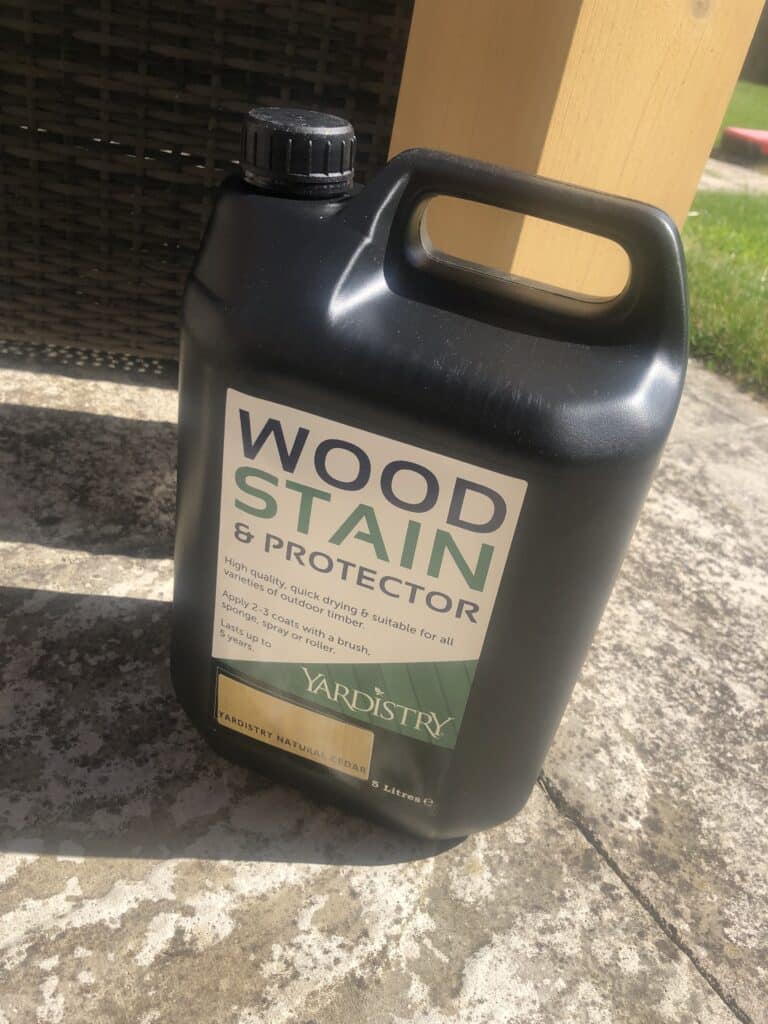 Black plastic container of Yardistry Wood Stain & Protector placed on concrete outdoors, with wooden post and green grass in the background.
