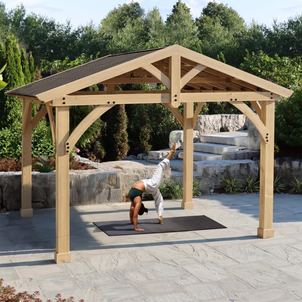 Person performing a handstand on a black mat under a wooden pergola in a landscaped garden.
