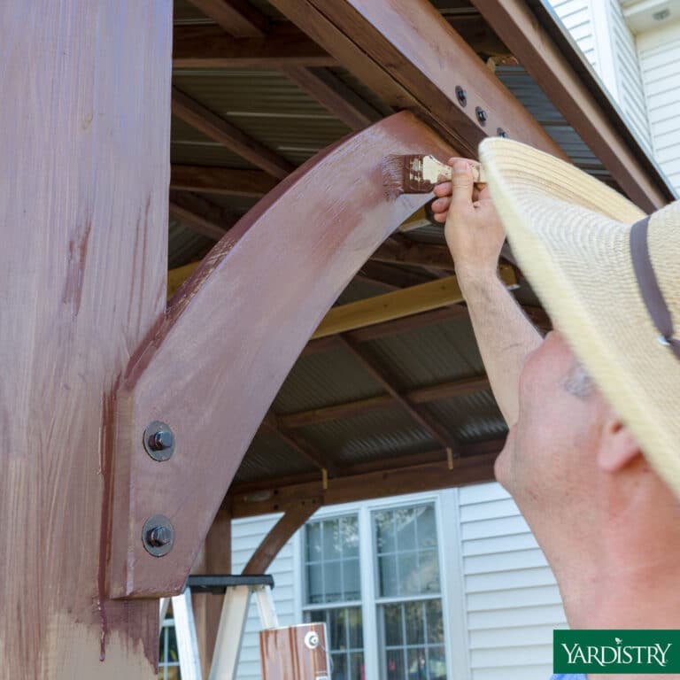 Person applying brown wood stain to a curved beam on a pergola using a brush, with bolts, corrugated roof, ladder, and white house siding visible in the background.