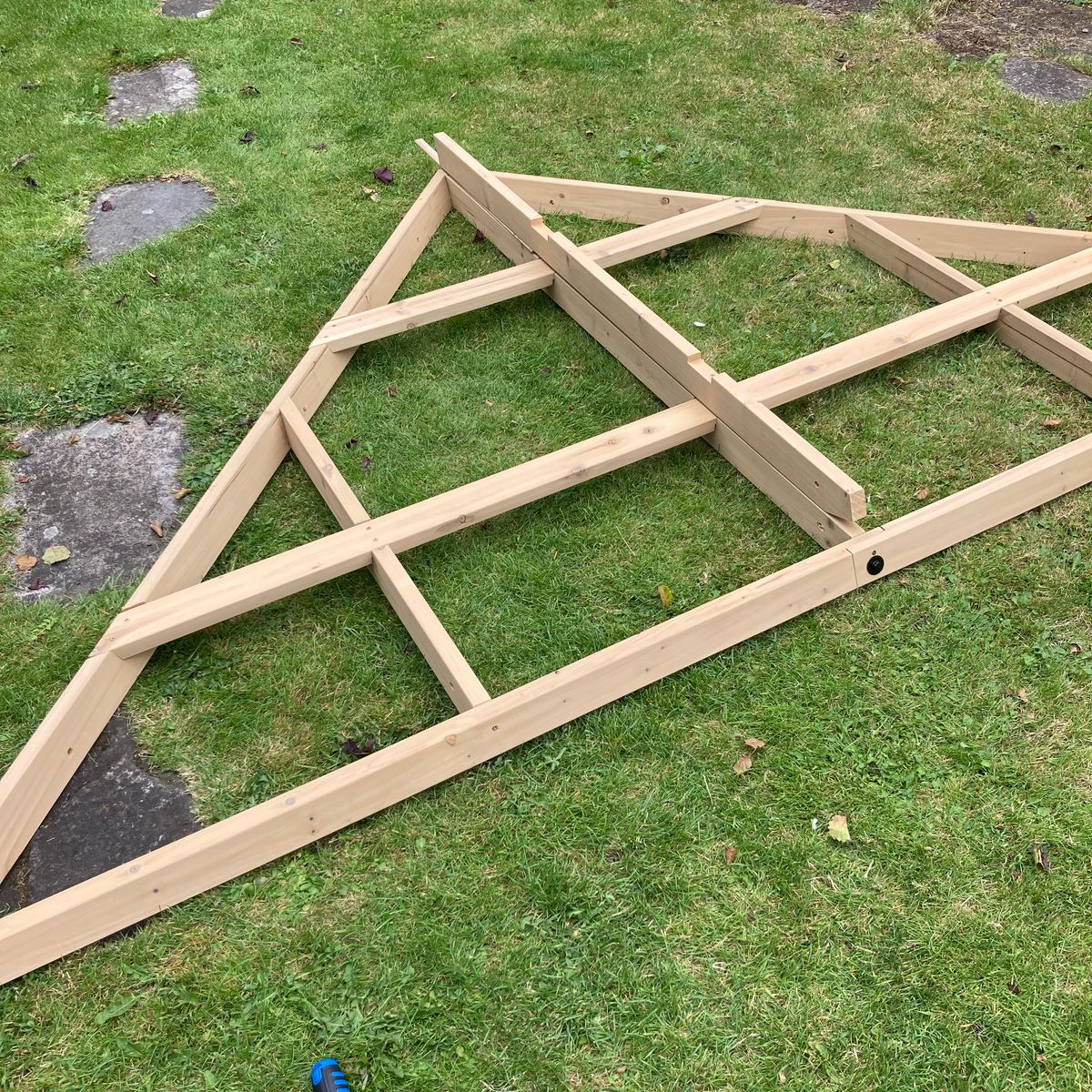 Triangular wooden frame with diagonal and horizontal supports lying on a grassy lawn, surrounded by stepping stones and garden greenery.