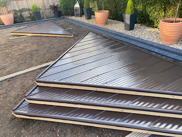 Triangular corrugated metal panels with beige trim laid out on soil in a landscaped garden area, surrounded by wooden planks, stone borders, and potted plants.
