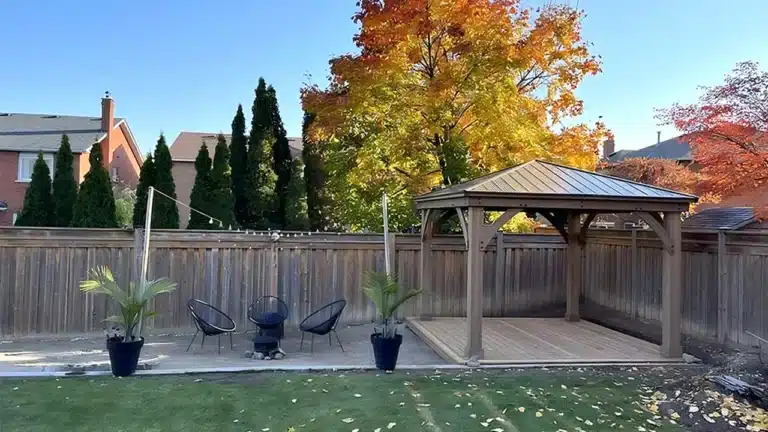 Wooden gazebo in a sunlit autumn garden with patio seating, potted plants, and vibrant fall foliage