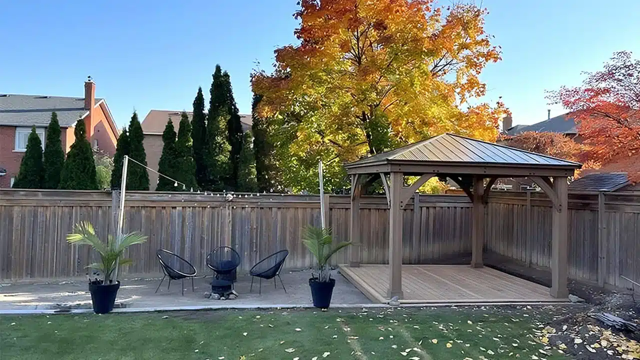 Wooden gazebo in a sunlit autumn garden with patio seating, potted plants, and vibrant fall foliage