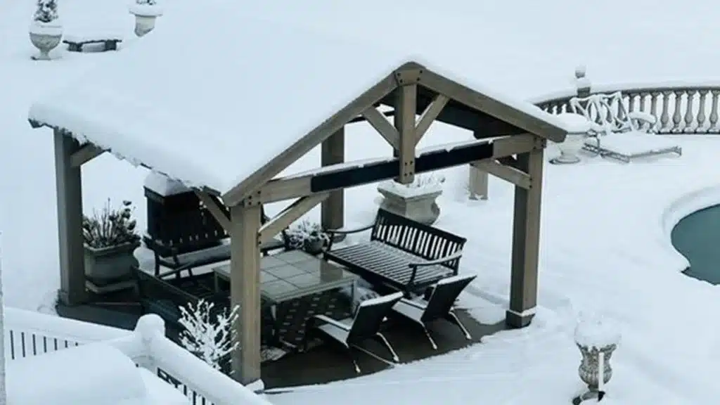 This image captures an elevated view of a wooden outdoor pavilion and its surrounding patio, completely covered in a fresh blanket of snow. The scene is dominated by white, evoking a peaceful, cold winter atmosphere.

The pavilion is the central feature, constructed with robust, dark-stained wooden posts and beams supporting a steeply pitched, gable-style roof. The roof is heavily laden with snow, highlighting its protective function. The interior structure features dark bracing timbers.

Beneath the roof, the space is furnished with dark-colored outdoor furniture, all slightly dusted or covered with snow. Visible items include a square or rectangular dining table, several modern lounge chairs with black sling fabric, and a porch swing or bench swing hanging from one of the cross beams. A few dark planters with dormant or snow-covered plants are also situated within the shelter.

The pavilion stands on a paved or decked area, which blends seamlessly into the surrounding snow-covered patio. The background reveals a formal, balustrade-lined stone patio, where classic stone urns or planters, also snow-capped, are placed. To the right, the curved edge of a swimming pool, partially covered in snow, adds a distinct feature to the landscape. The overall picture contrasts the sturdy, dark wooden structure with the soft, pristine white of the winter snow.