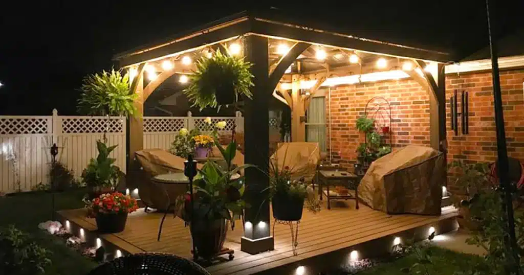 This image depicts a cozy, fully illuminated outdoor living space centered around a wooden gazebo set against a dark night sky.

The gazebo has a dark, hard-top roof and a sturdy frame of natural or stained wood posts and beams, featuring large arched supports. Warm, exposed-filament string lights are hung inside the structure, casting a bright, inviting glow. The structure sits atop a raised wooden deck with a light brown finish. The edge of the deck is outlined with small, white or warm-toned ground lights, highlighting the perimeter.

The space is furnished with outdoor seating, although most pieces are covered with light brown or tan protective covers, suggesting the evening is either cool or the furniture is being preserved. A round table and several chairs are visible. The area is heavily decorated with greenery and potted plants, including large planters on the deck, hanging ferns from the gazebo beams, and colorful flowers near the front. A square, illuminated planter base sits centrally, adding another layer of ambient light.

The gazebo is positioned against a house wall made of red brick, which reflects the warm interior light. To the left, a low, white lattice privacy fence borders the yard, with more subtle lighting on the grass. The overall impression is a highly decorative, well-lit, and functional backyard retreat, despite the furniture being covered.