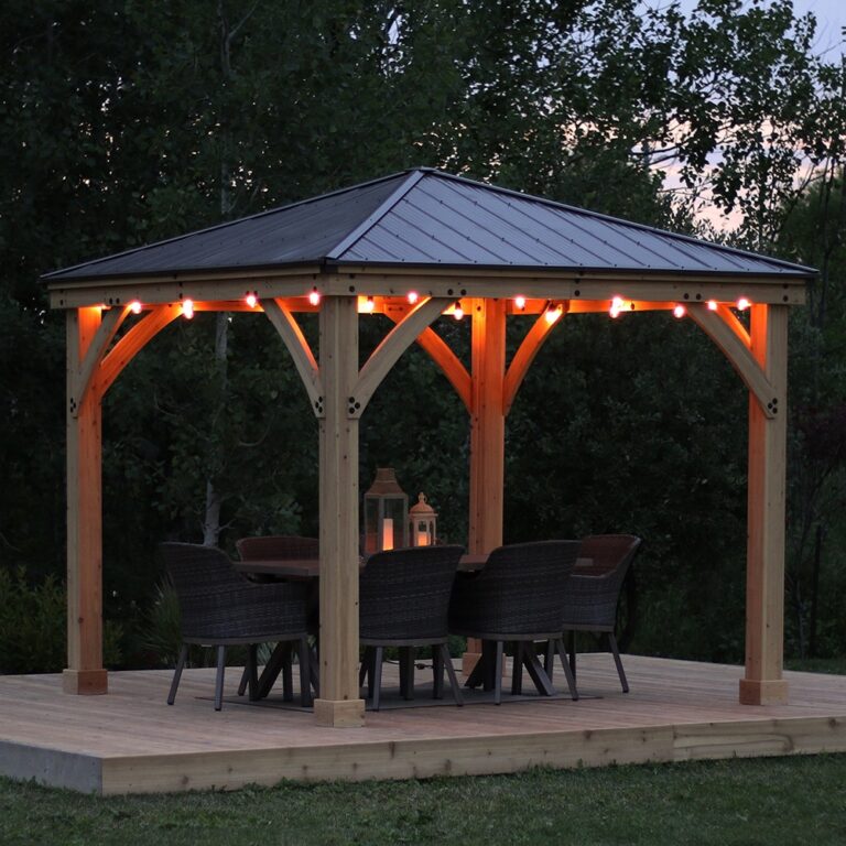 Wooden gazebo with metal roof and string lights on a raised platform, featuring a dining table with wicker chairs and lanterns, surrounded by trees and grass at dusk.