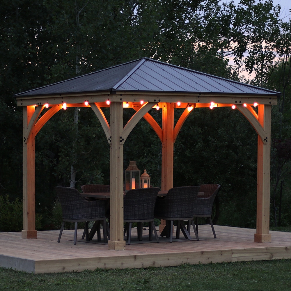 Wooden gazebo with metal roof and string lights on a raised platform, featuring a dining table with wicker chairs and lanterns, surrounded by trees and grass at dusk.