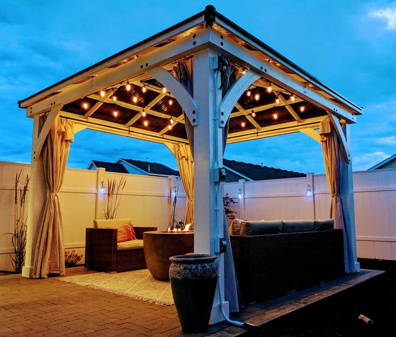 This image captures a beautifully designed outdoor pavilion, or gazebo, set up as a comfortable and inviting evening retreat. The structure features a substantial white wooden frame with striking arched supports connecting the posts to the roof, giving it a sturdy, elegant look. The roof itself has a rustic, corrugated metal underside, from which a series of warm, exposed filament string lights are suspended, casting a golden, ambient glow over the seating area below.

Cream or beige-colored outdoor curtains are draped and tied back to the posts on all sides, adding a sense of enclosure, privacy, and soft texture. The floor is paved with dark brick or stone pavers.

Inside the pavilion, two distinct seating areas are visible: on the left, there's a pair of woven wicker or rattan armchairs situated on a light-colored patterned area rug. In the center of this arrangement is a large, cylindrical fire pit or outdoor table with a concrete-like finish. A glimpse of a small fire or decorative flame suggests it's in use. On the right, a dark-colored wicker outdoor sofa provides additional seating.

The entire area is enclosed by a white privacy fence. The dusk setting, with a deep indigo-blue sky, contrasts beautifully with the bright interior lighting of the pavilion and the softer blue accent lighting visible on the fence posts and behind some ornamental grasses, creating a very serene and cozy atmosphere for outdoor relaxation or entertaining.