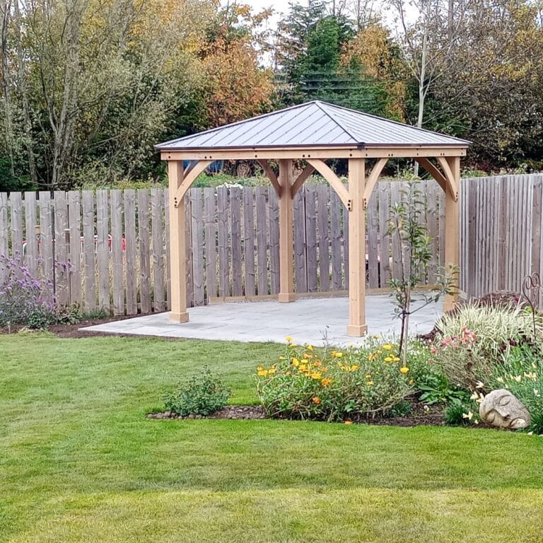 Wooden gazebo with a metal roof on a concrete pad, surrounded by a privacy fence, flower beds, and autumn trees in a landscaped backyard.