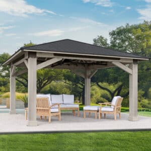 Wooden outdoor gazebo with dark pitched roof and cushioned seating, set on a paved surface near grass, trees, and a pool.