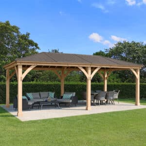 Large wooden pavilion with dark metal roof and cross-bracing, featuring cushioned seating and dining area on a concrete pad in a landscaped garden.
