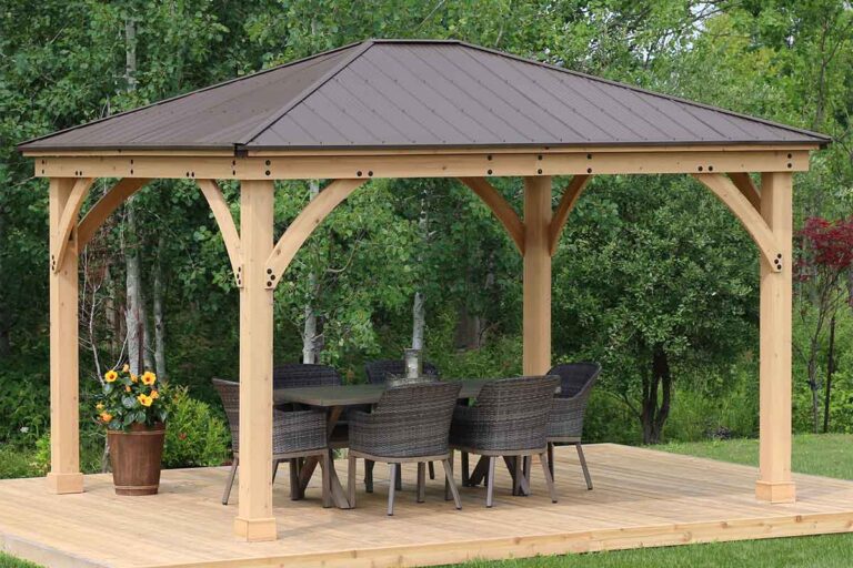 Wooden gazebo with dark metal roof and wicker dining set, placed on a wooden deck in a lush garden with trees and foliage.