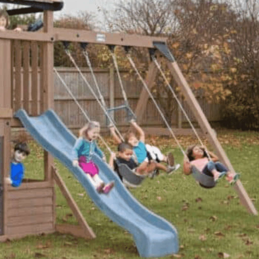 Children playing on a wooden outdoor playset with swings, a slide, and a small enclosed area, set in a grassy garden with autumn trees and a wooden fence in the background.