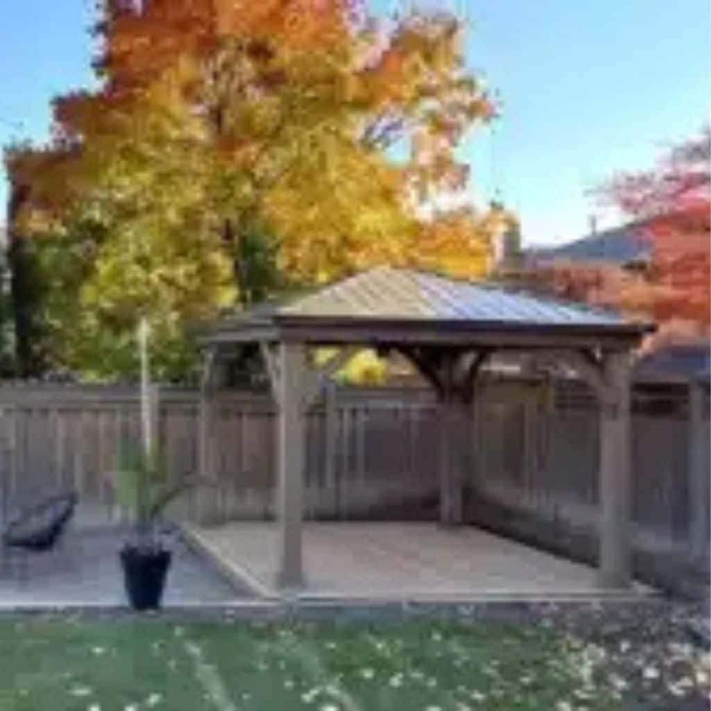 Wooden gazebo in a sunlit autumn garden with patio seating, potted plants, and vibrant fall foliage.