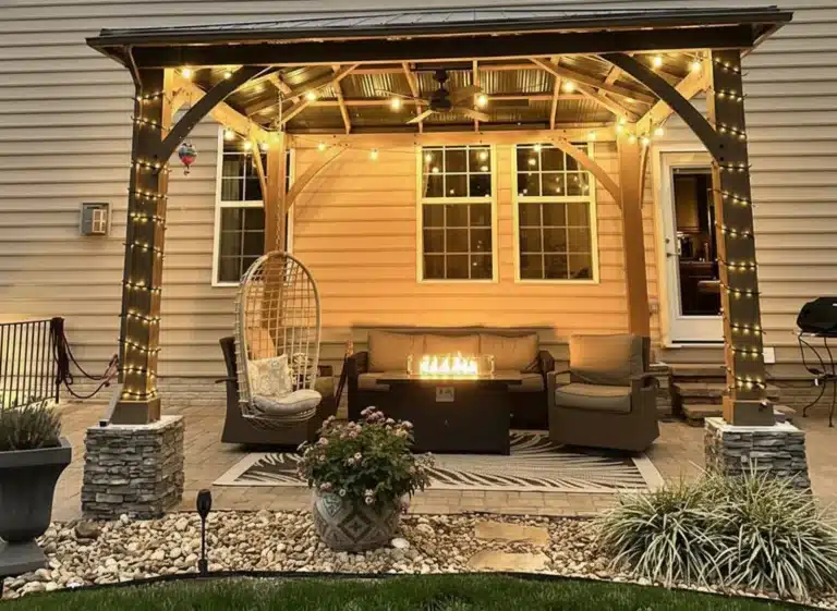 An outdoor patio at night featuring a large wooden gazebo with a dark metal roof. The gazebo is illuminated by warm string lights wrapped around its pillars and hanging from the rafters, as well as an integrated ceiling fan with lights. Underneath, there is a cozy seating area with a wicker sofa and armchair, a hanging egg chair, and a rectangular fire pit table with a glass wind guard. The area is set on a stone paver patio with a patterned rug, surrounded by decorative rock landscaping, potted plants, and the side of a beige-sided house with two lit windows in the background.